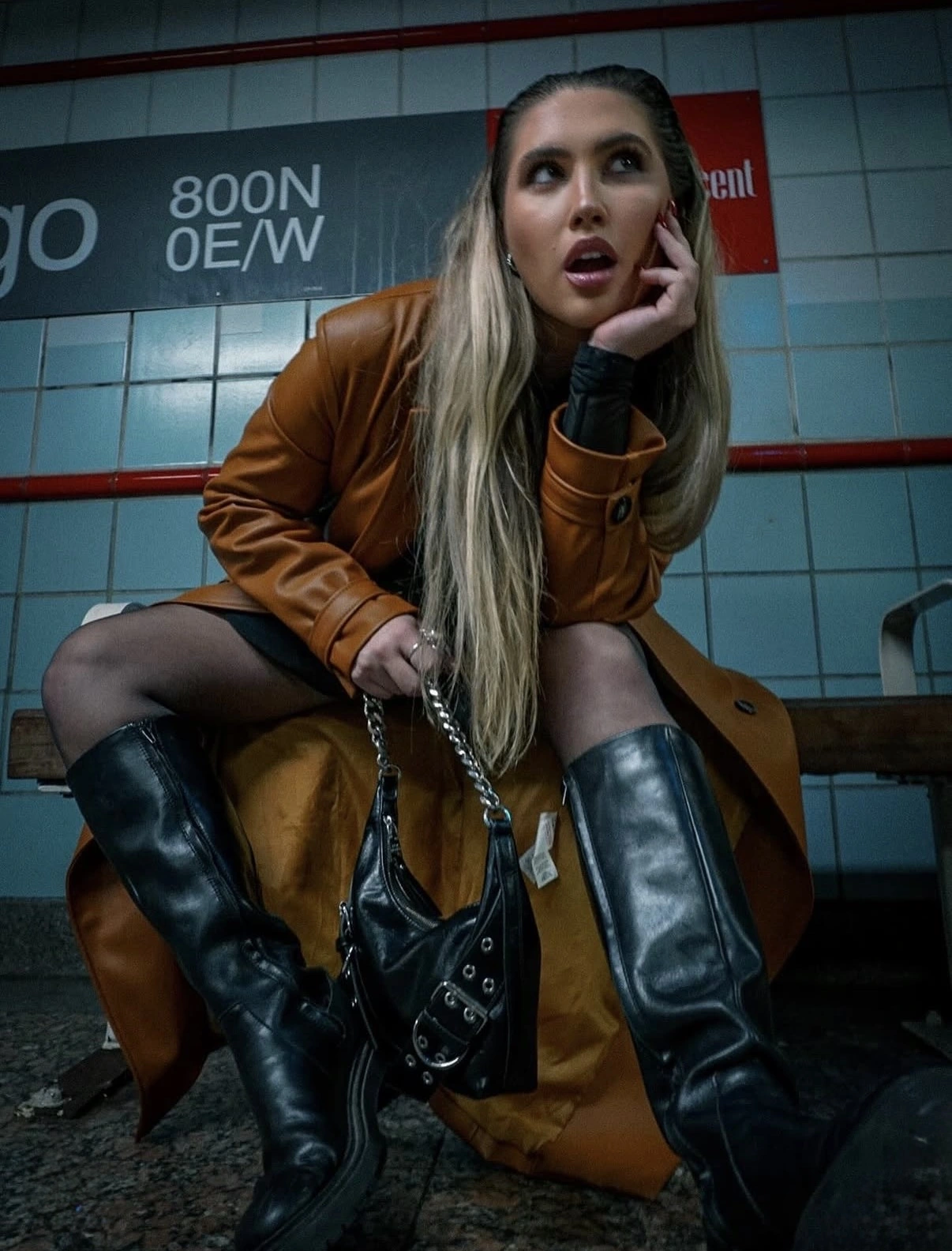 Woman in a brown coat and tall black boots seated at a train station