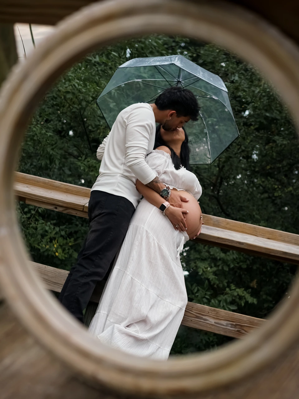 Expectant couple kissing beneath a clear umbrella, framed by a circular mirror