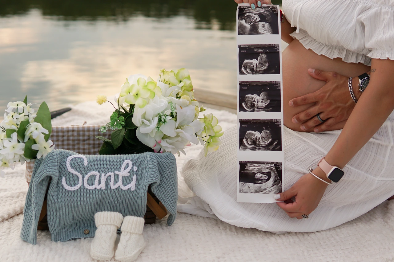 Hands holding ultrasound photos beside a pregnant belly, flowers and a blue Santi sweater