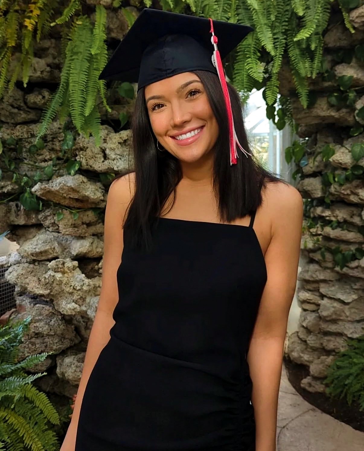 Graduate in a black cap and dress smiling among ferns and stone walls in a conservatory